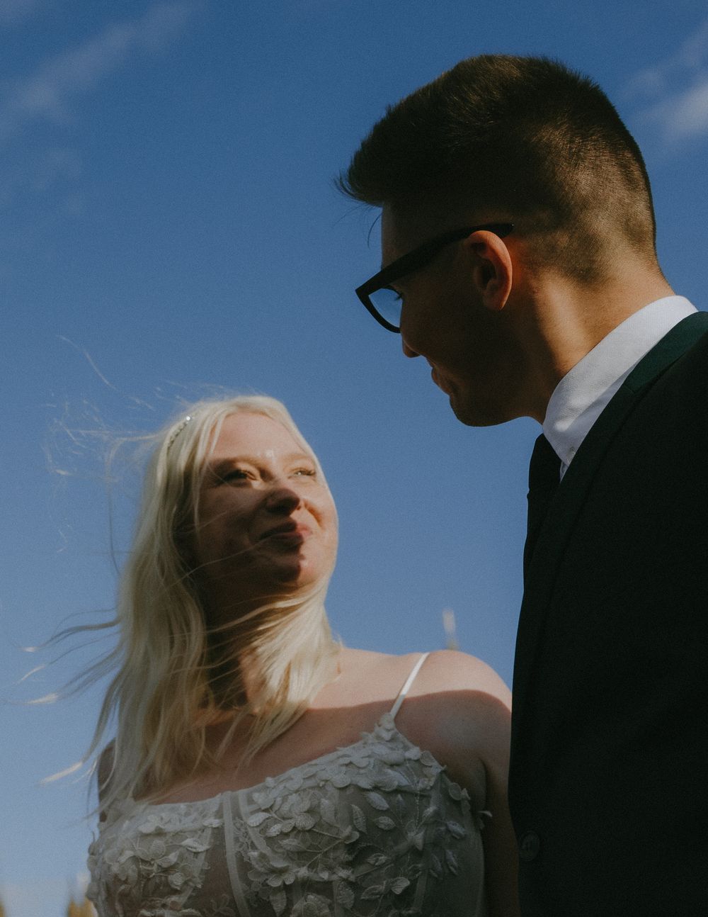 A romantic wedding photo of a couple against a blue sky, with her white dress and veil flowing in the breeze.