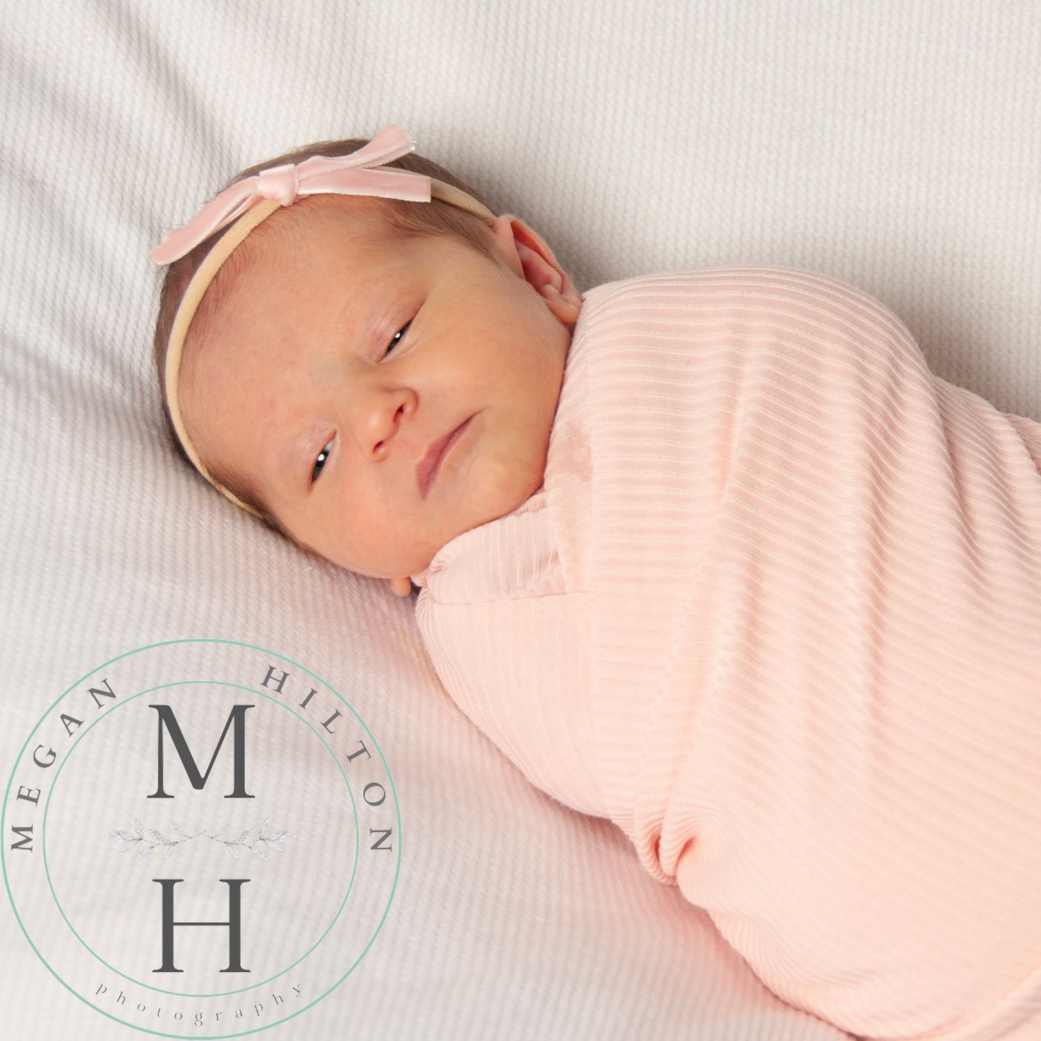 An almost sleeping newborn baby swaddled in light pink with matching headband rests on white bedding.