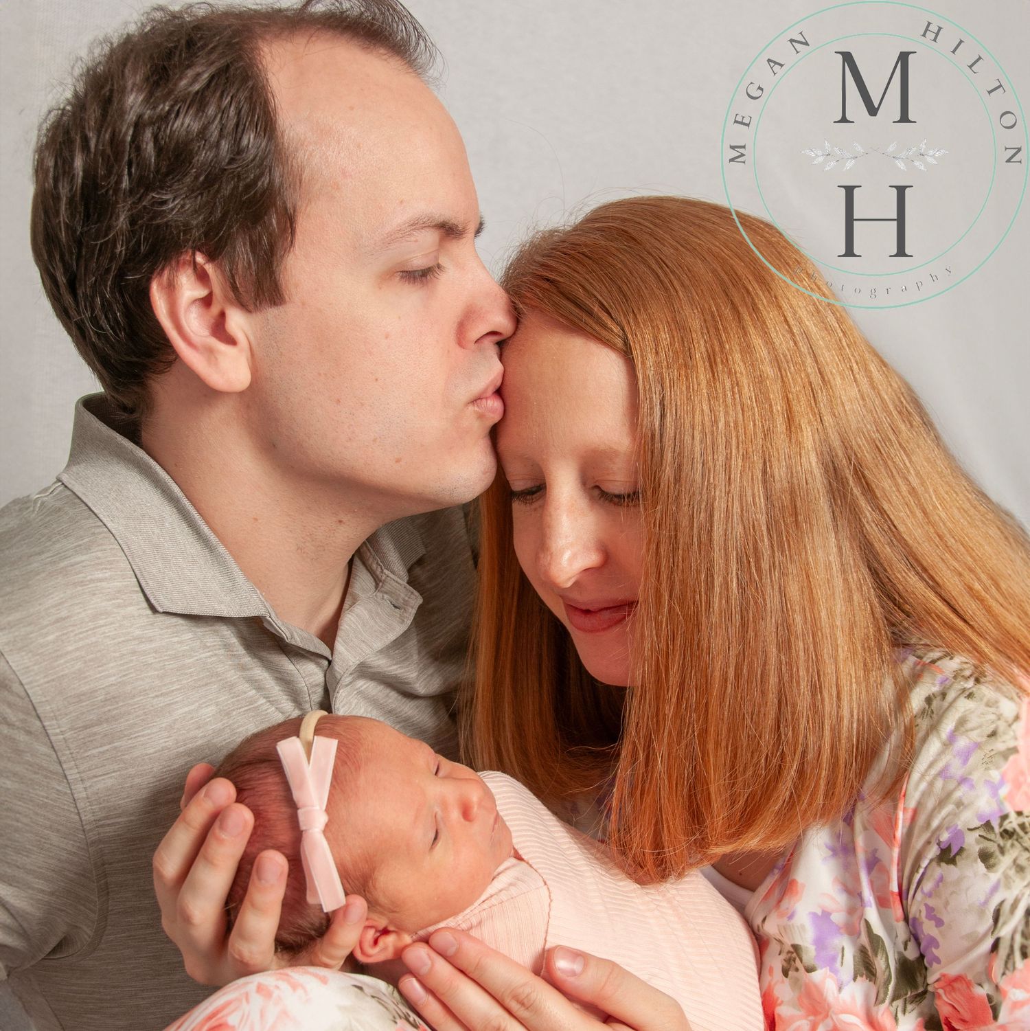 A family moment as a dad gently kisses the mom while holding their newborn baby in a soft studio setting.