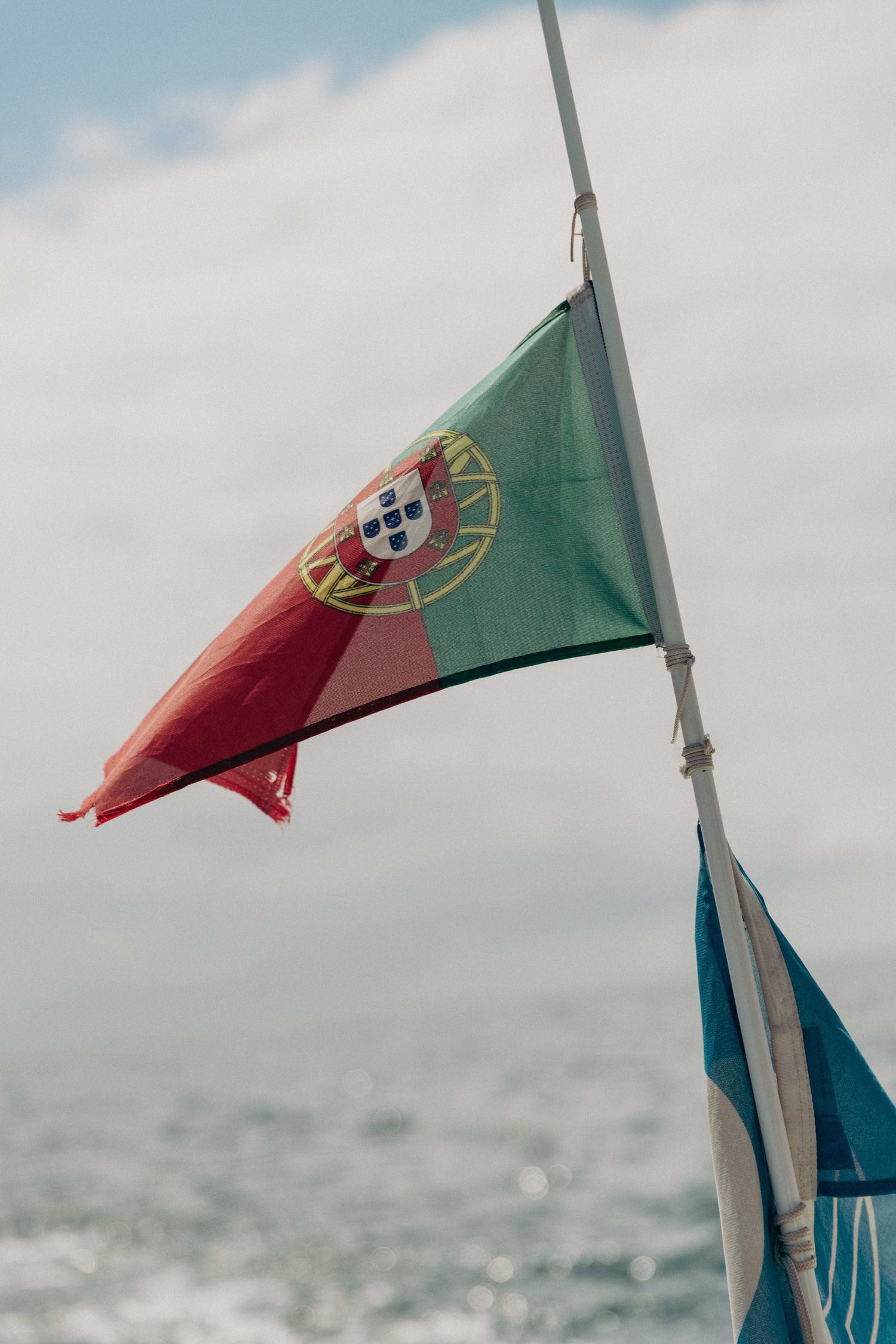 Portuguese flag waves in the wind against a cloudy sky and ocean backdrop with another flag visible below.