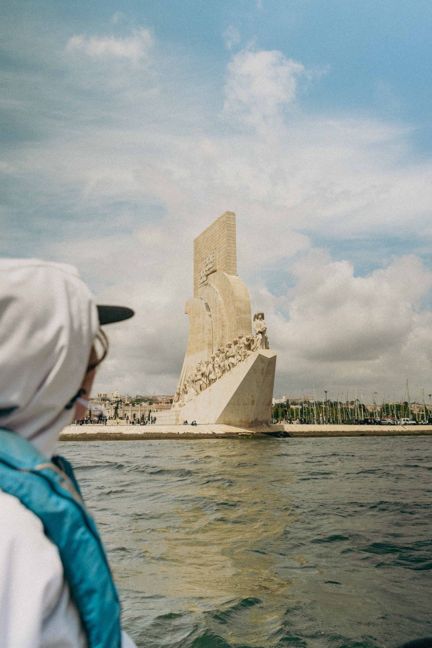 View from water of the Monument to the Discoveries in Lisbon with cloudy sky and waterfront backdrop.