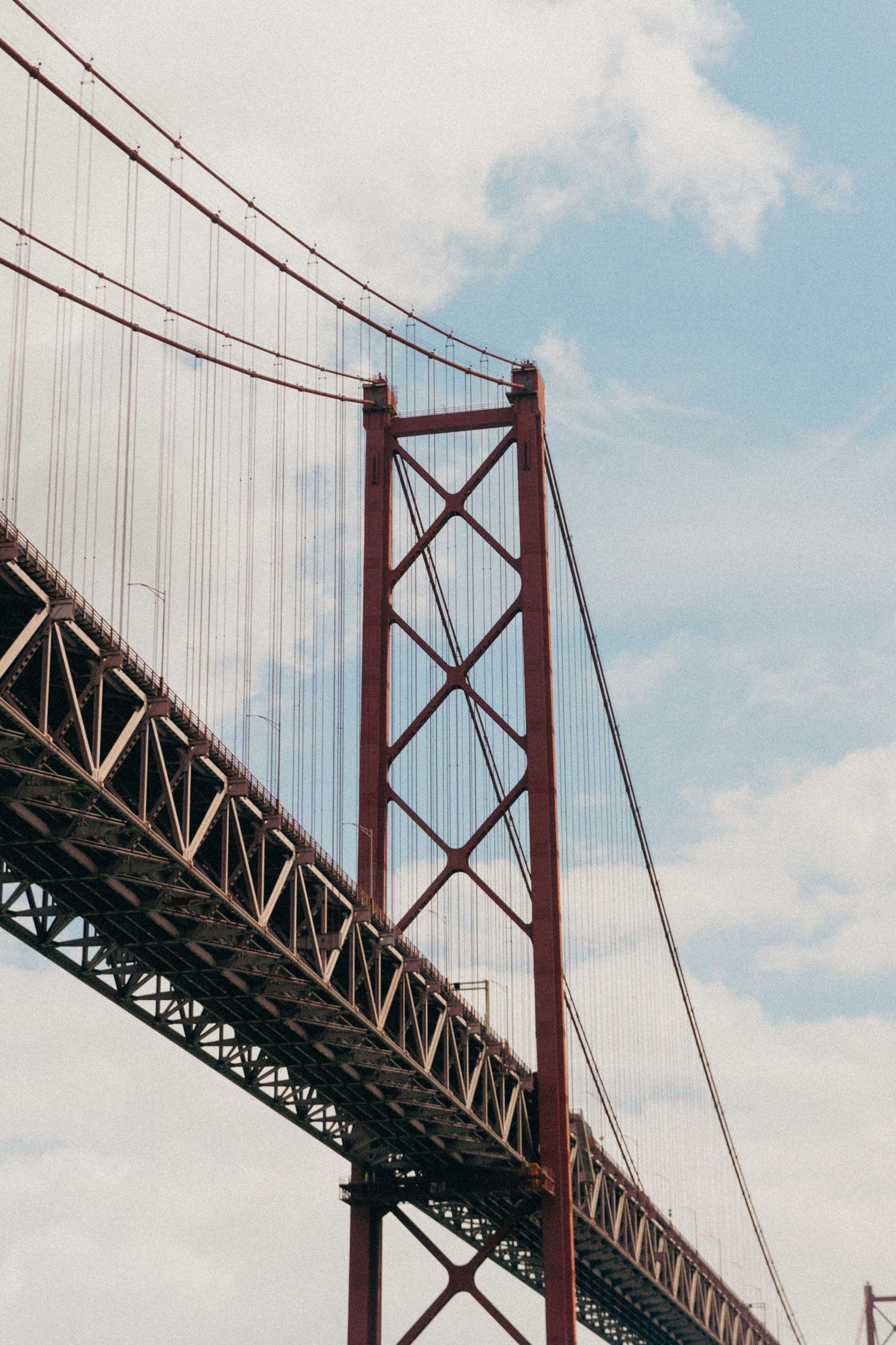 Looking up at the iconic red suspension towers and cables of the 25 de Abril Bridge in Lisbon against a blue sky.