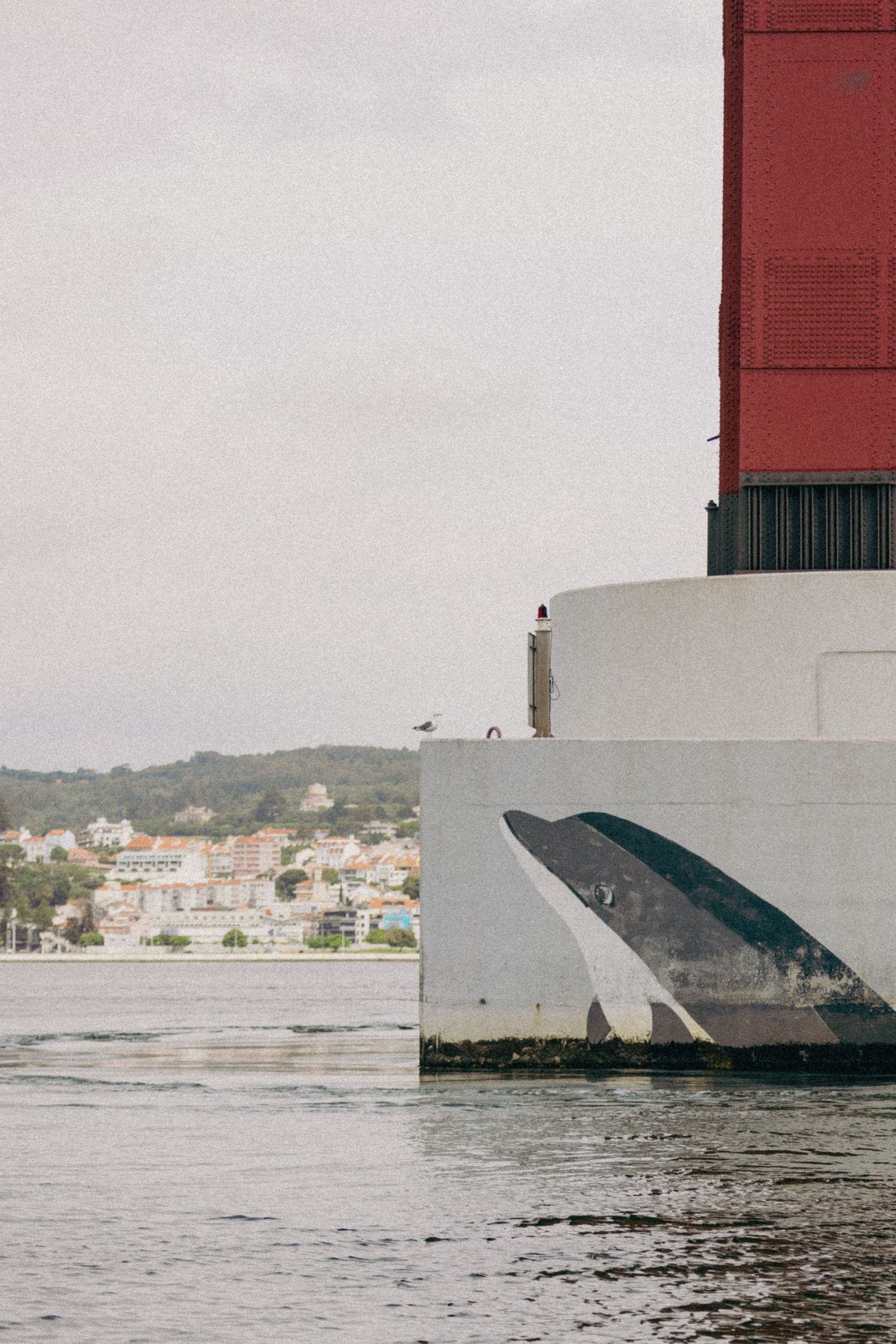 Small figure stands atop a white and red ship hull against a coastal town backdrop over choppy waters.