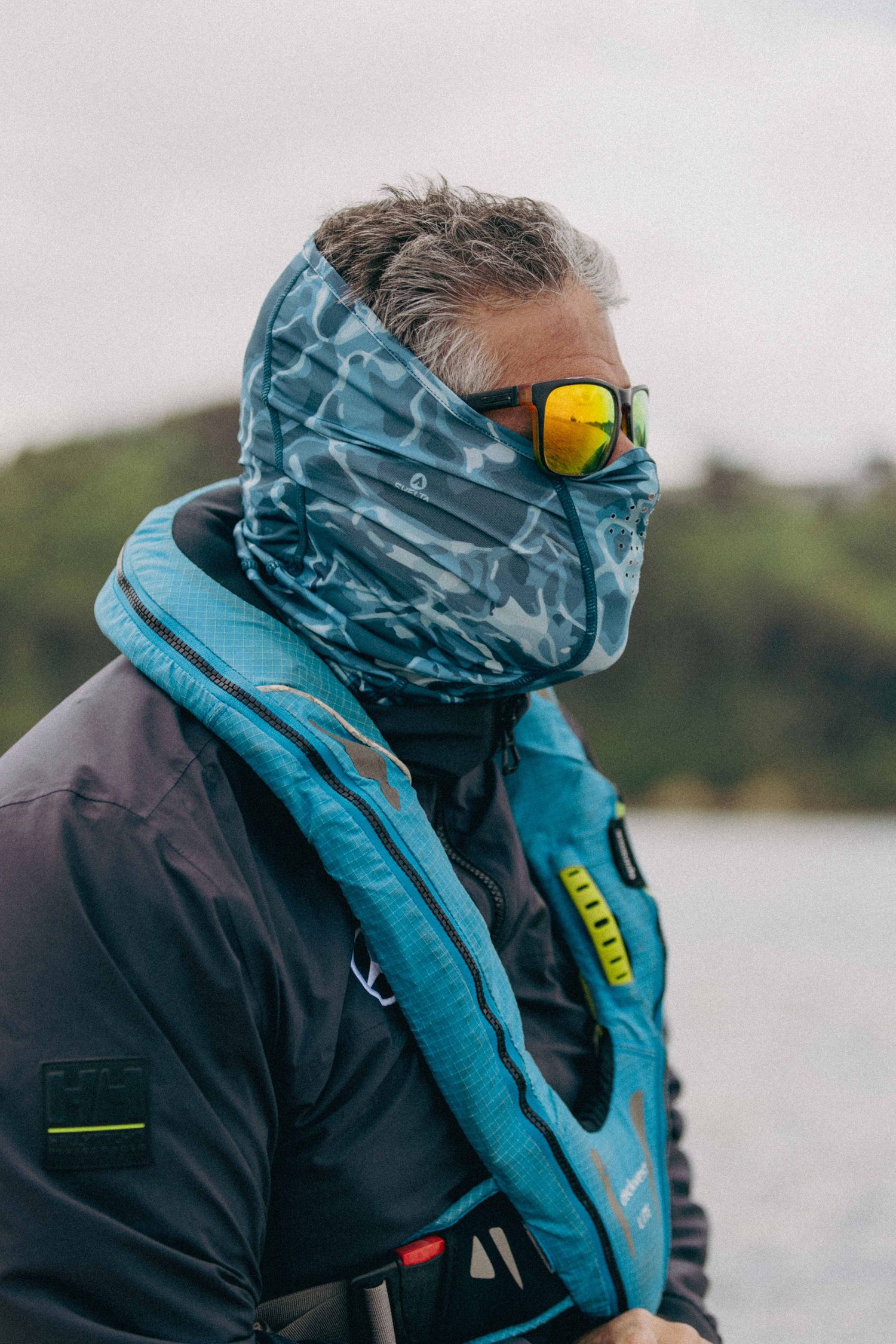 Person wearing blue neck gaiter and yellow mirrored sunglasses stands by water wearing dark outdoor gear.
