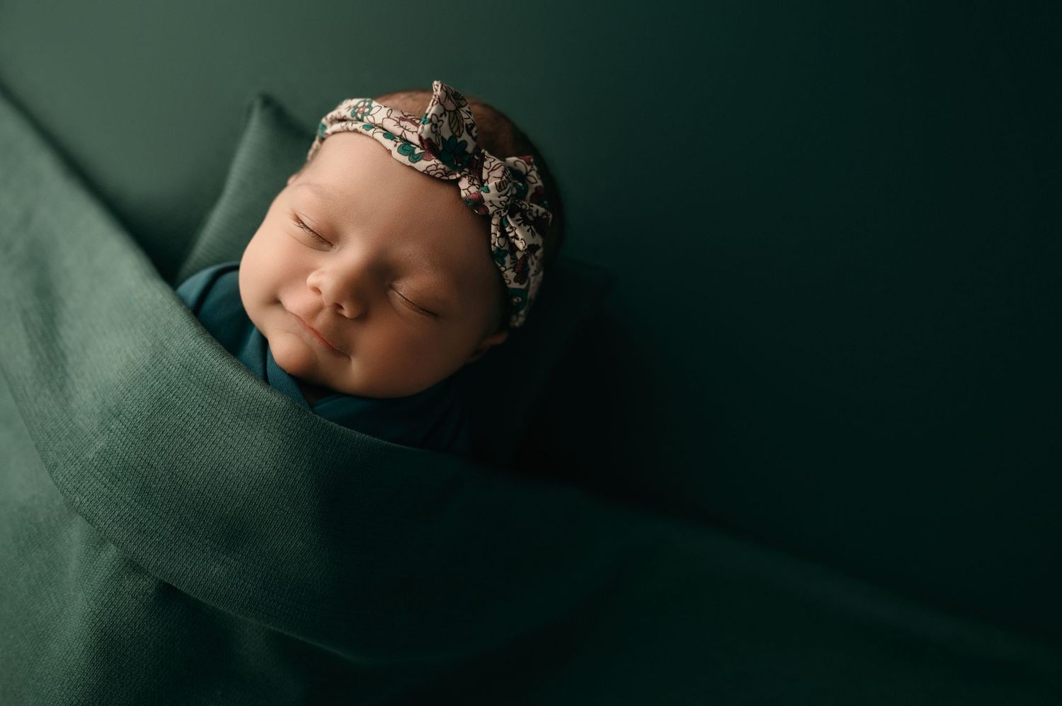 Sleeping newborn wearing floral headband nestles peacefully in emerald green wrap during professional photo session.