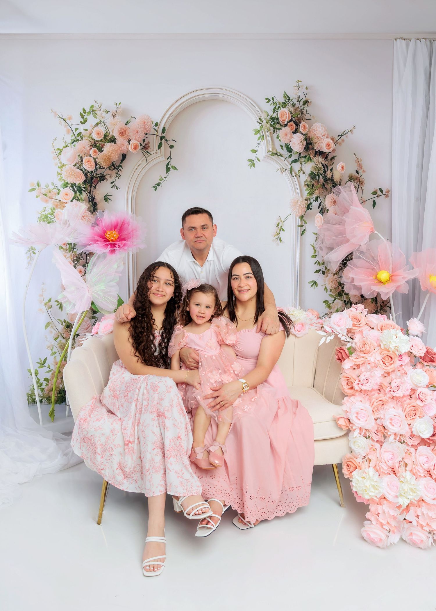 Family of four posing on a cream sofa surrounded by pink floral arch decorations in a white studio.