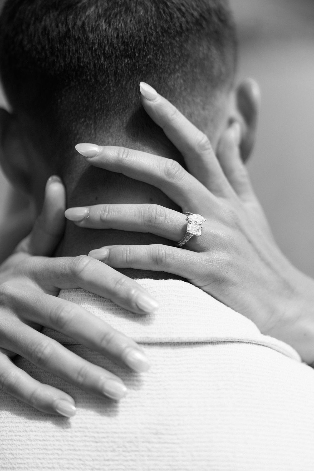 Black and white photo of a woman's hand with diamond engagement ring resting on a man's neck.