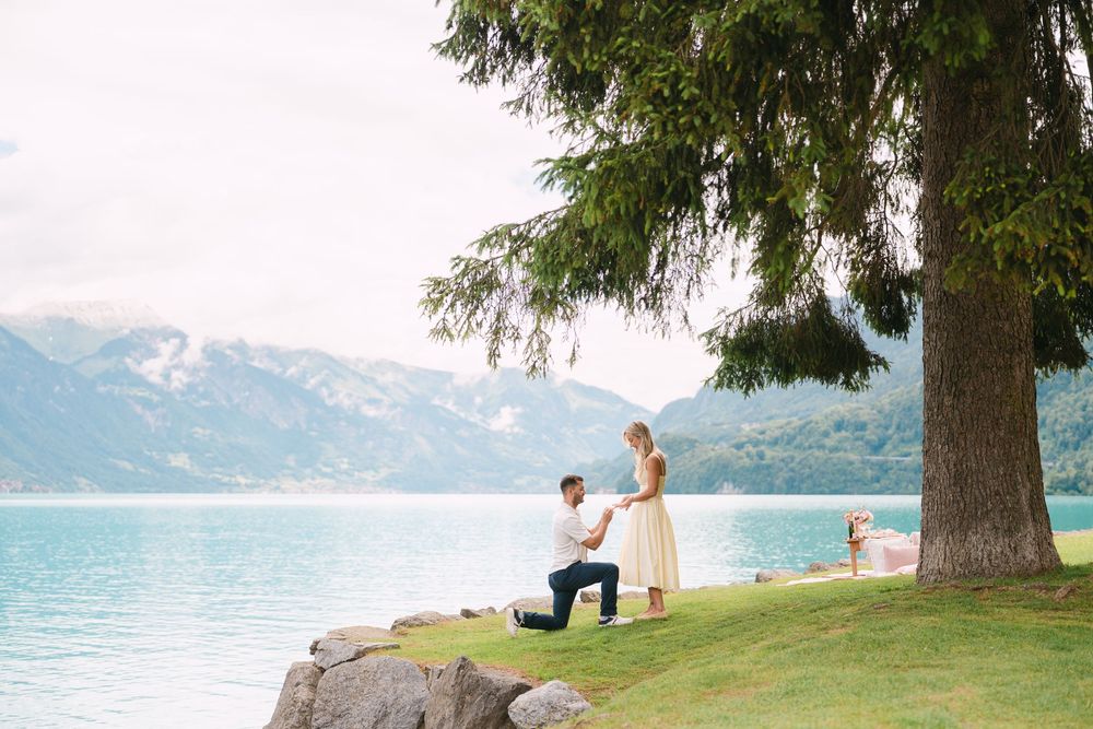 Man proposes on one knee to woman by turquoise lake with mountain backdrop under large tree.
