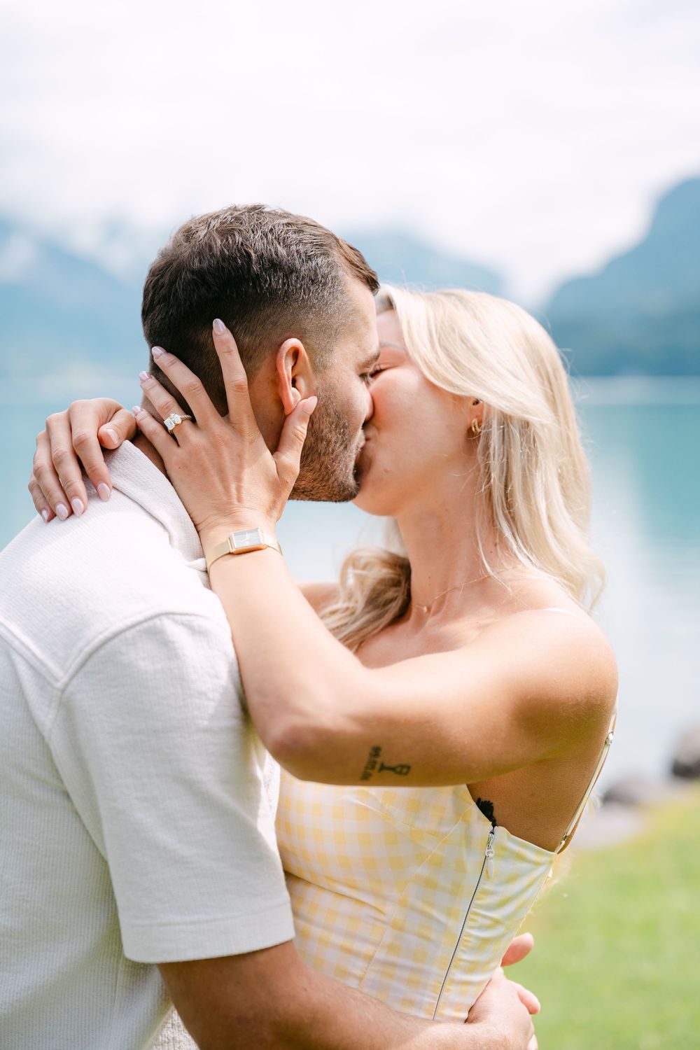 Couple sharing a romantic kiss by a scenic mountain lake, woman in yellow dress with ring visible.