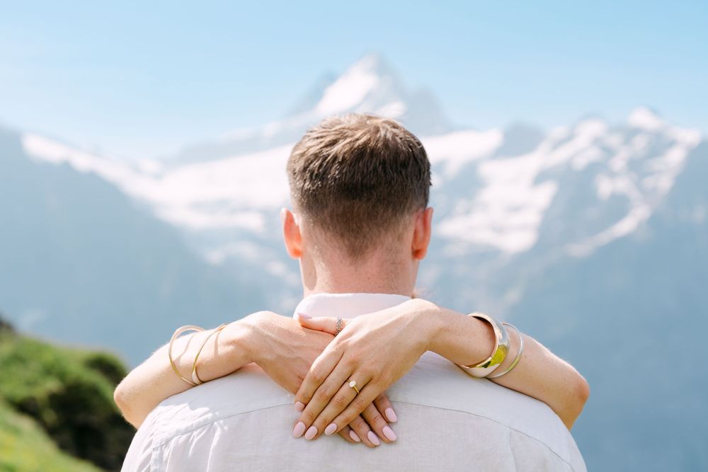 Couple embracing with scenic snow-capped mountain backdrop, woman's hands with gold bracelets resting on man's shoulders.