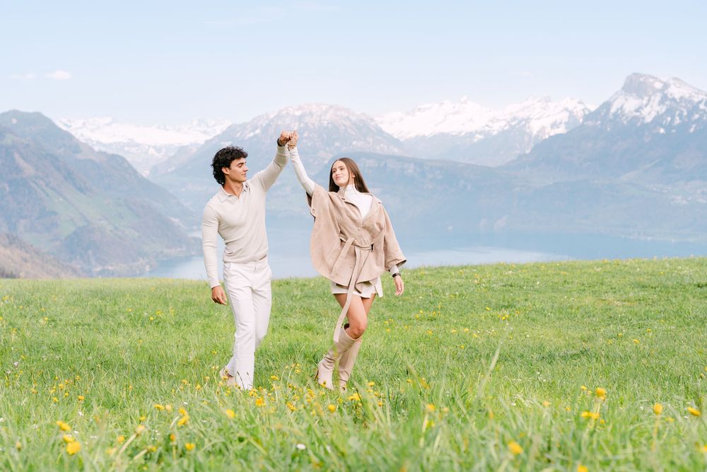 Couple dancing joyfully in a green meadow with snow-capped mountains and a lake in the background.