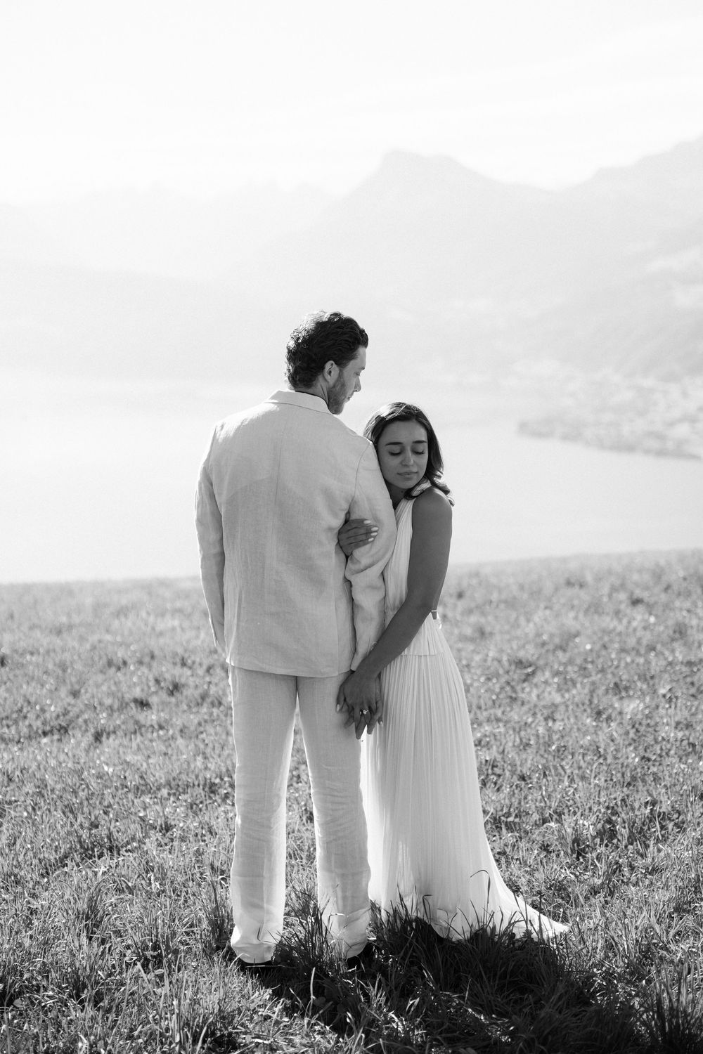 Black and white photo of a couple embracing in a field with a lake and mountains in the background.