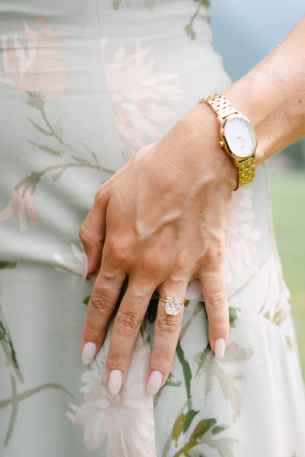 Close-up of a woman's hand wearing a gold diamond ring and gold watch, resting on a floral dress.