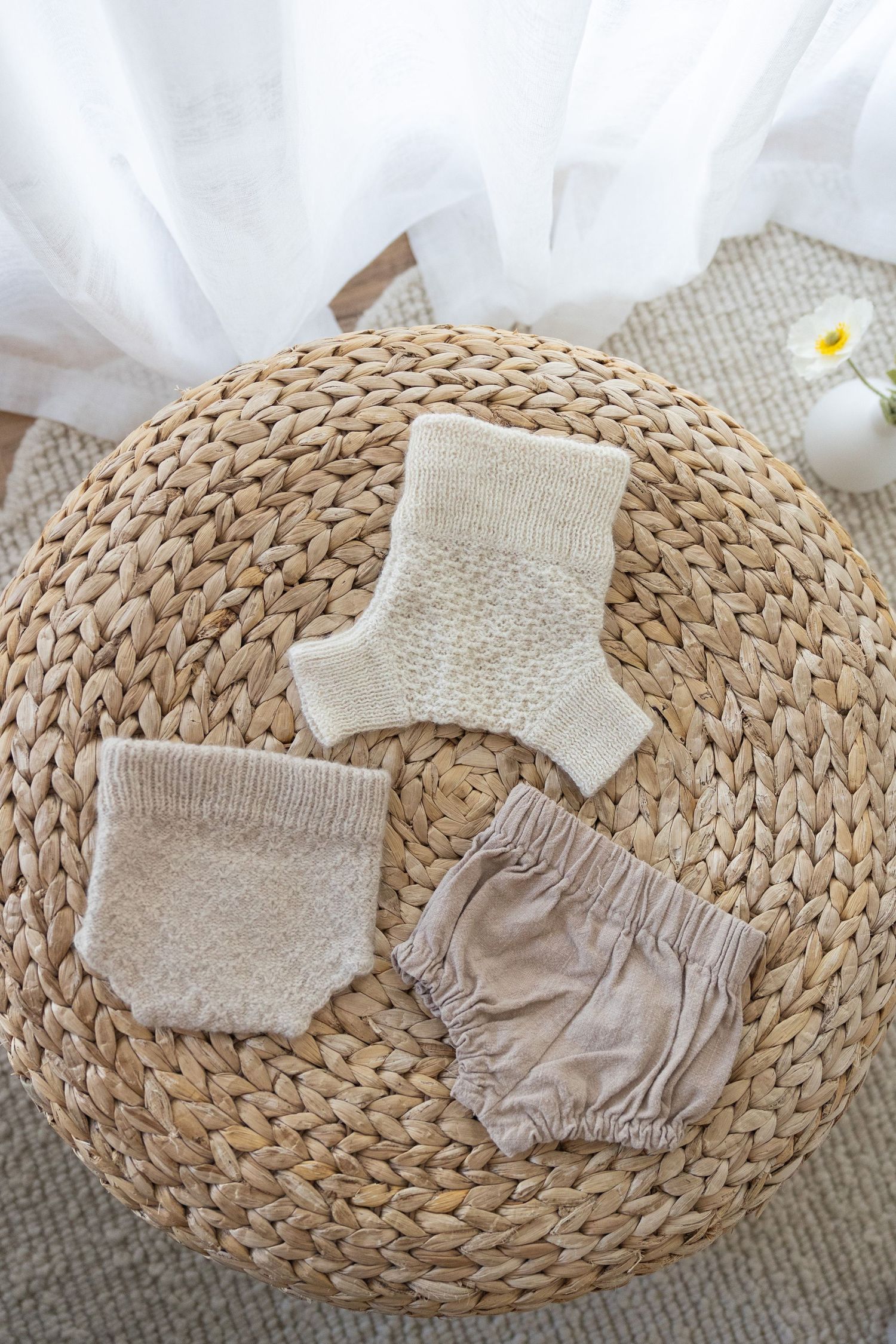 Baby clothes and socks arranged on a round woven rattan mat with neutral color tones and soft textures.