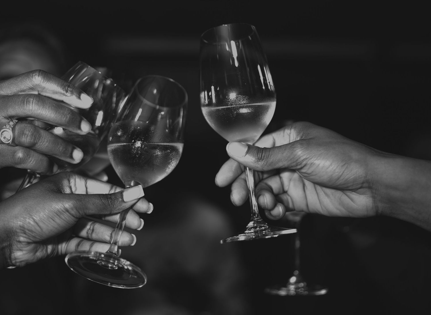Three hands raise wine glasses together in a celebratory toast against a dark background in black and white.