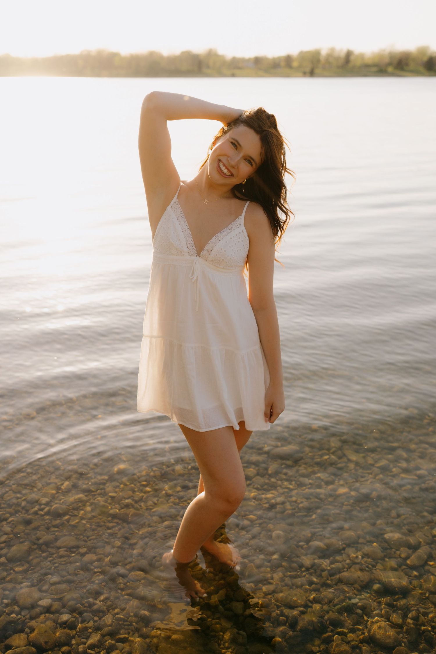 A person in a white sundress poses at the water's edge during golden hour sunset on a rocky beach.