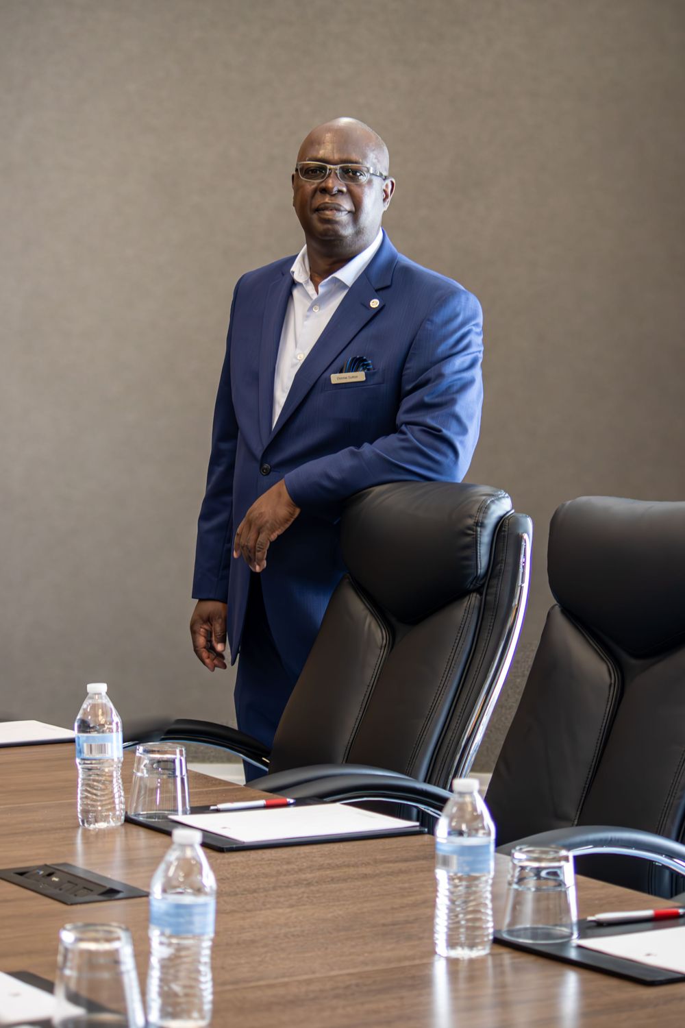 Professional man in blue suit standing confidently at a corporate conference table with water bottles and chairs.