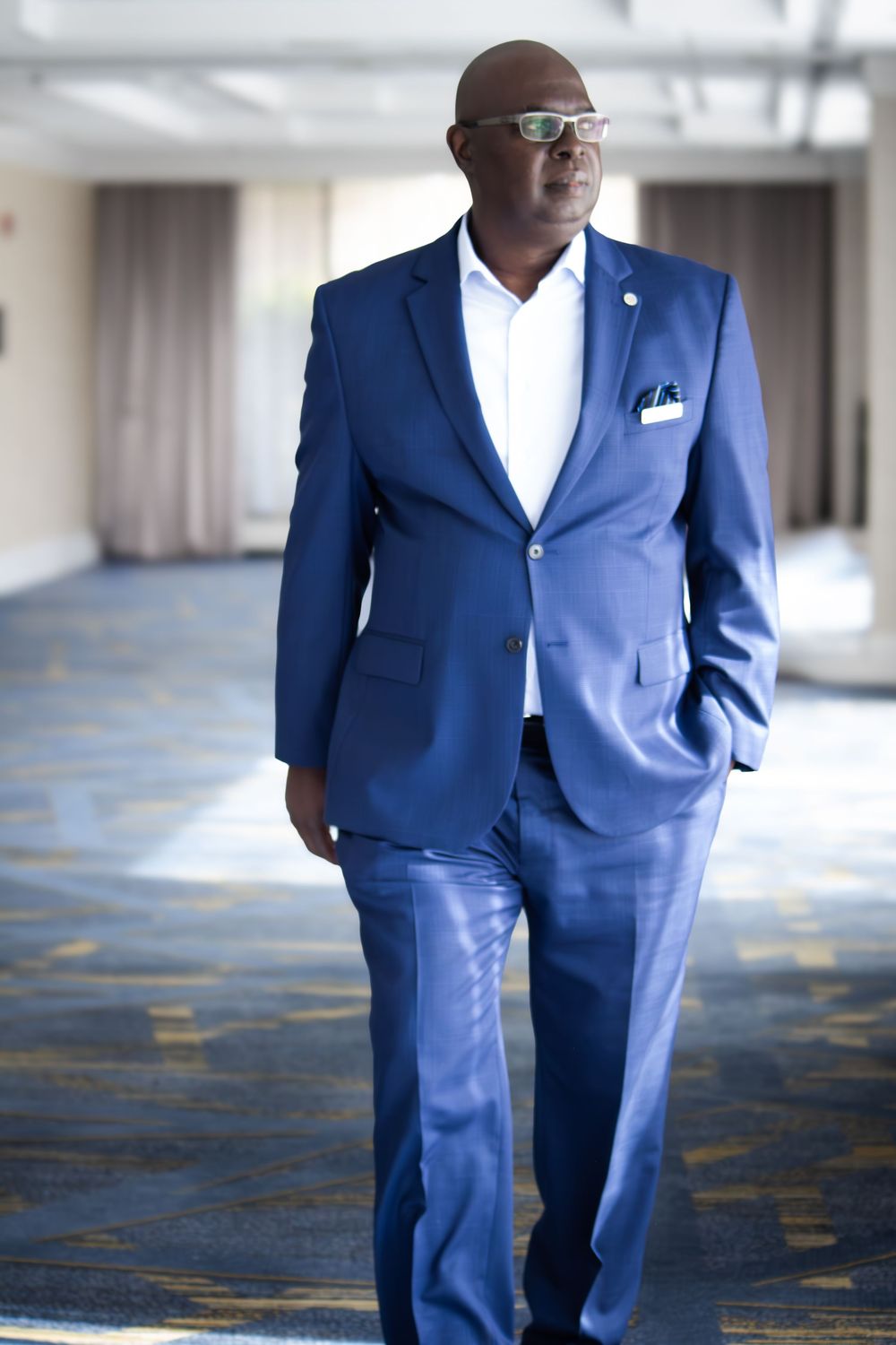 A confident bald man in a blue suit and glasses walks through a modern hallway.