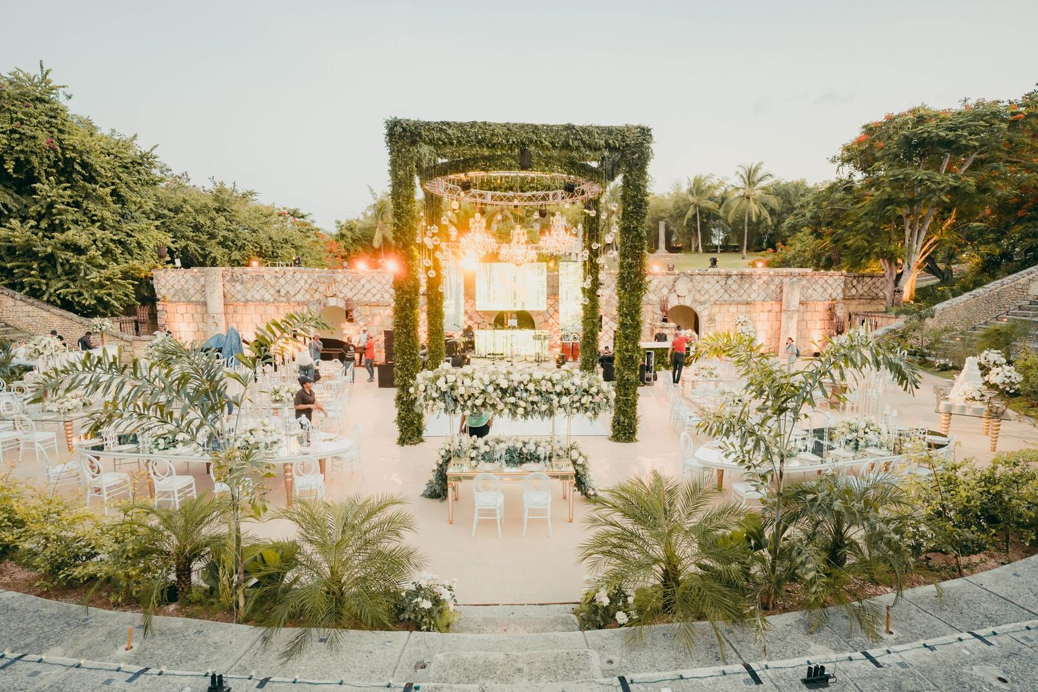 Elegant outdoor wedding ceremony venue with palm trees and floral arch lit by golden sunlight at sunset.