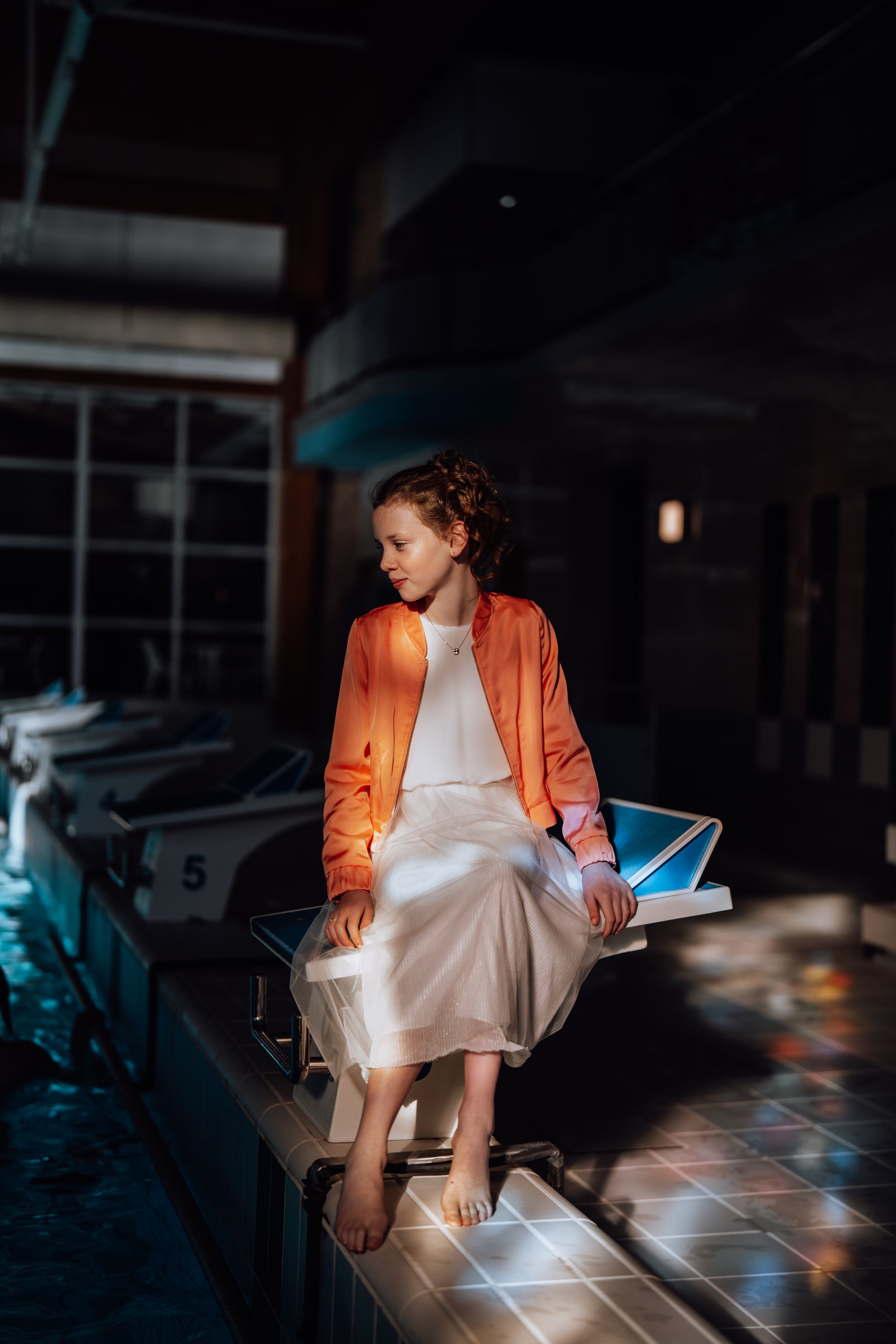 Figuur met oranje jas en witte jurk zit blootsvoets op betegelde richel in stemmige binnenverlichting met filmische sfeer.