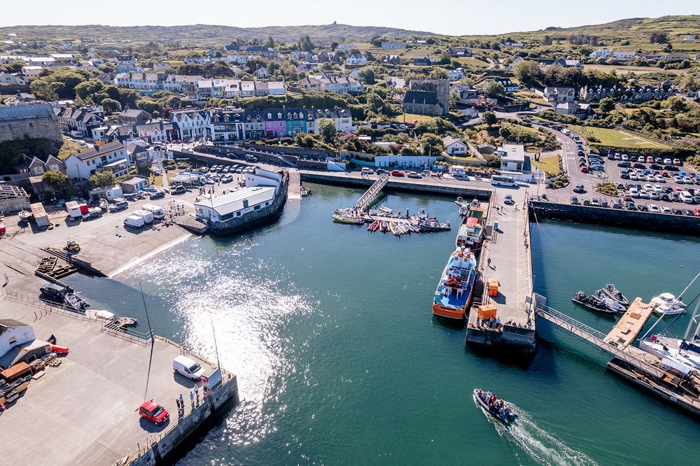 Aerial panoramic view of a coastal harbor with boats docked along the pier and surrounding hillside landscape.