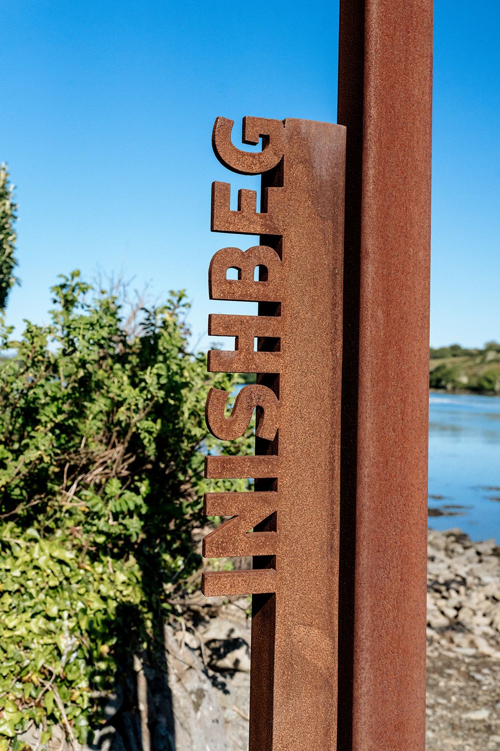 Rusty metal post with 'Inspire' text vertically mounted near a lakeside with greenery and water in the background.