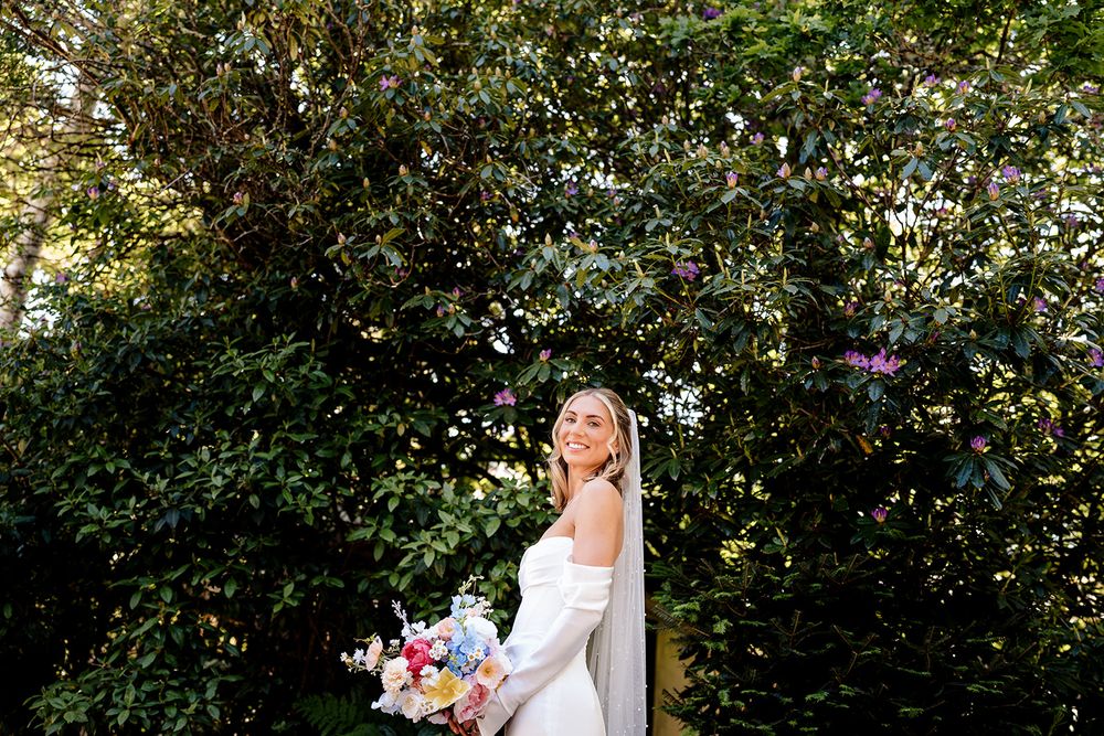 A bride in a white wedding dress holds a colorful bouquet while standing in front of lush green foliage.