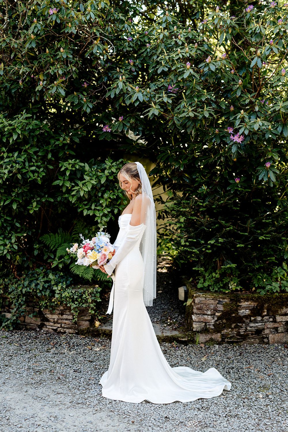 Classic white wedding gown with floral bouquet photographed against lush garden greenery on gravel path.