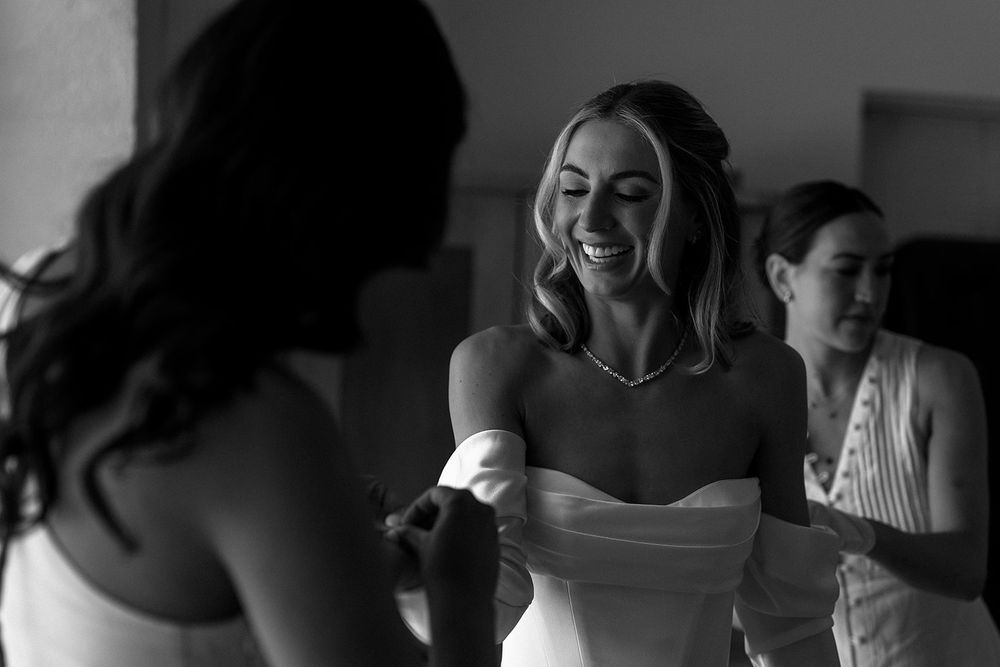 A joyful candid black and white moment showing a woman in an off-shoulder dress smiling during preparation.
