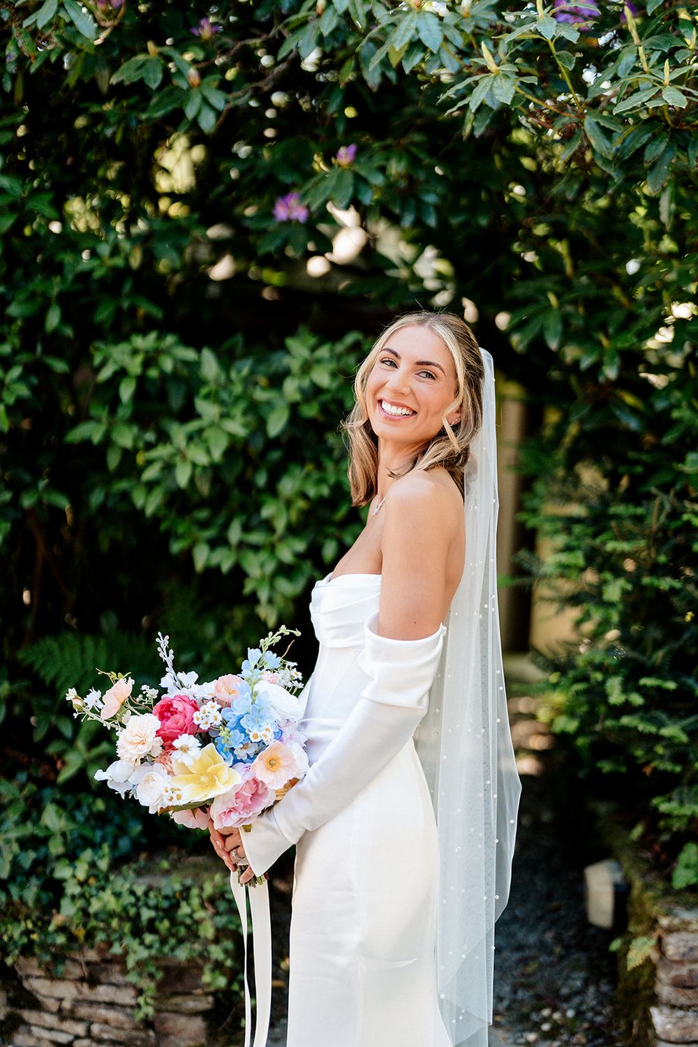 Bride in elegant white off-shoulder dress holds colorful bouquet of pink and blue flowers against lush garden background.