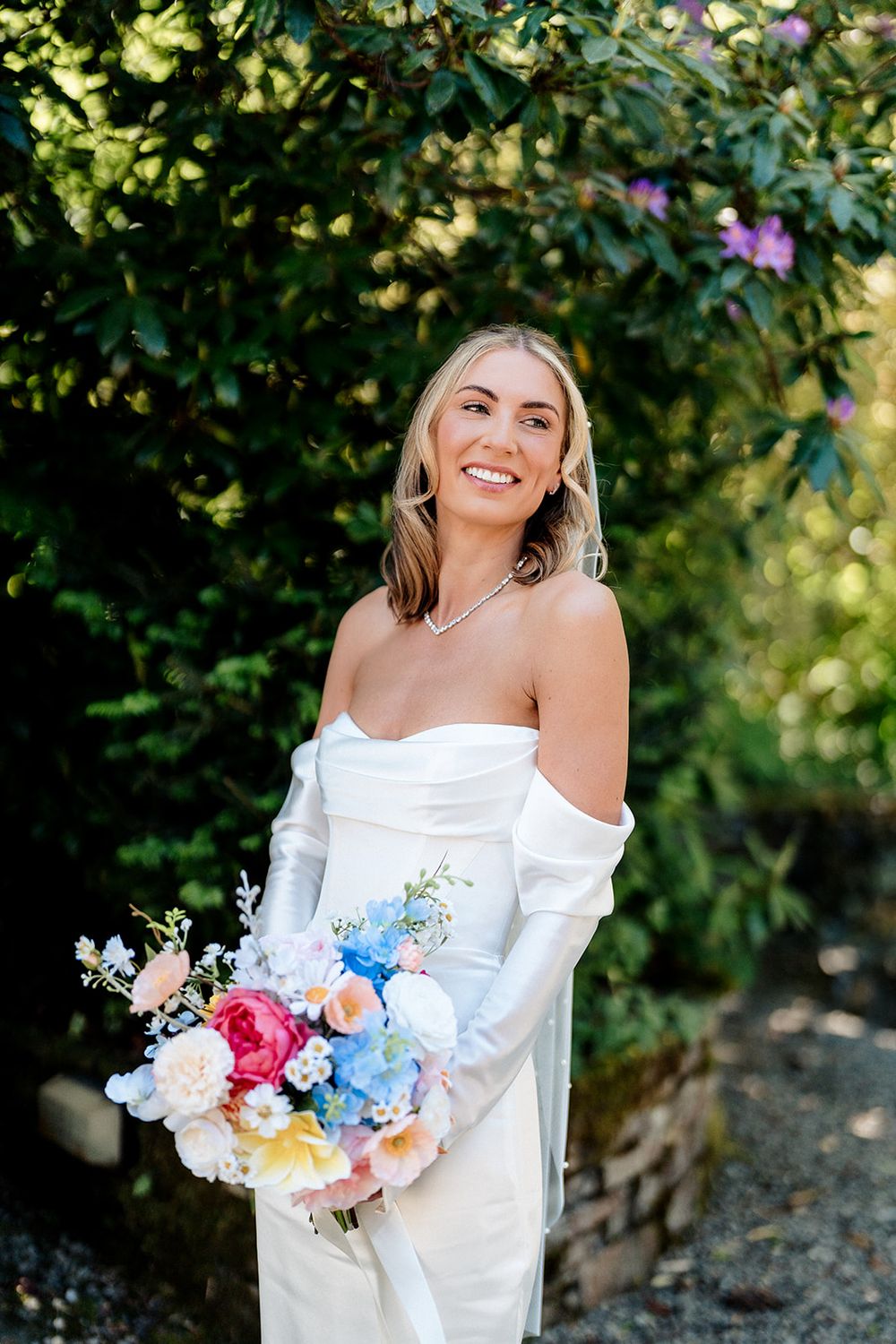 A bride in an elegant off-shoulder white dress holds a vibrant bouquet of pink, blue and white flowers in a garden setting.