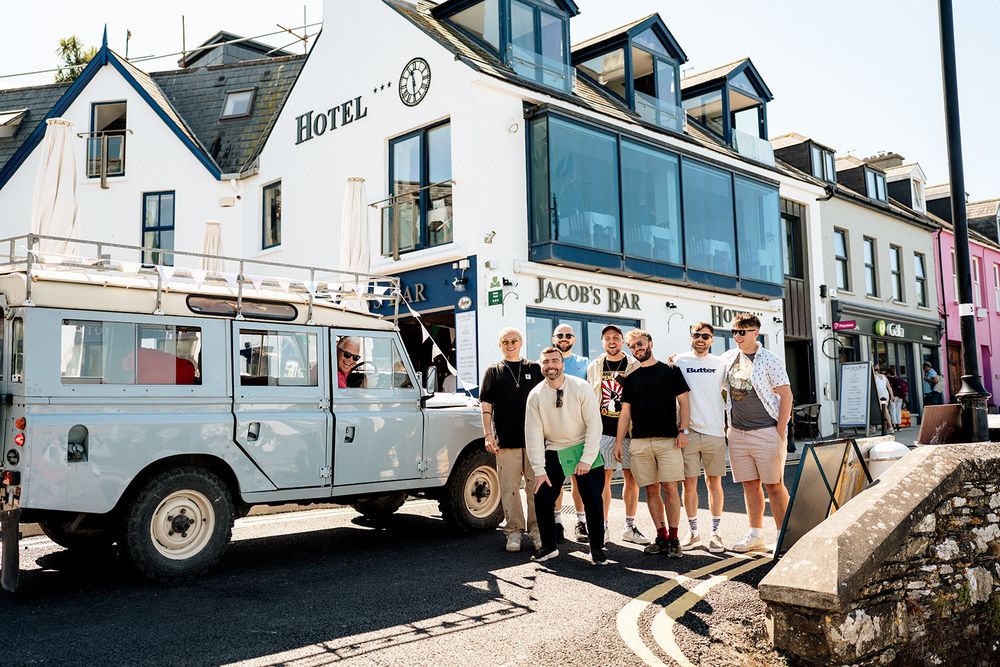 A vintage Land Rover parked outside a white hotel building with people gathered near Jacob's Bar in a coastal town.