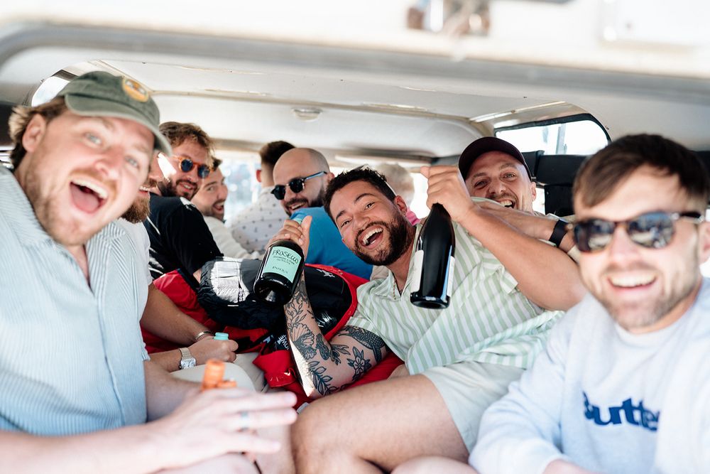 A group of friends laugh and hold beverages while sitting together in a vehicle for a casual gathering.
