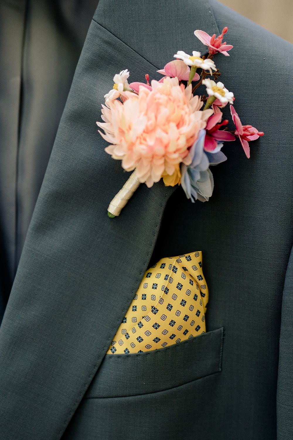 A peach-colored boutonniere with small flowers and a yellow patterned pocket square on a dark gray suit jacket.