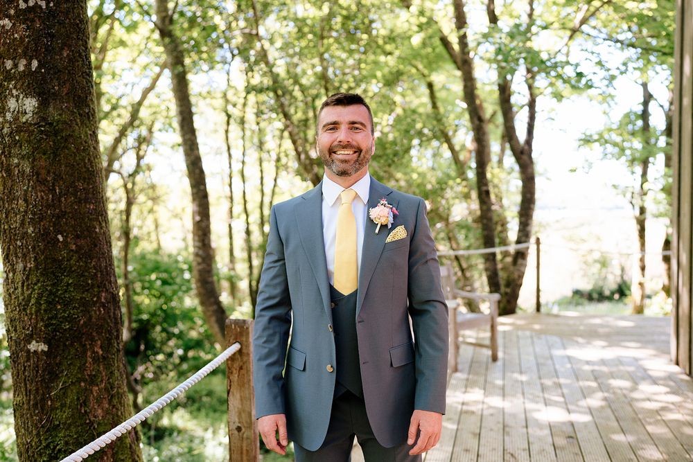 A groom in a charcoal suit with yellow tie stands smiling on a wooden walkway surrounded by trees.
