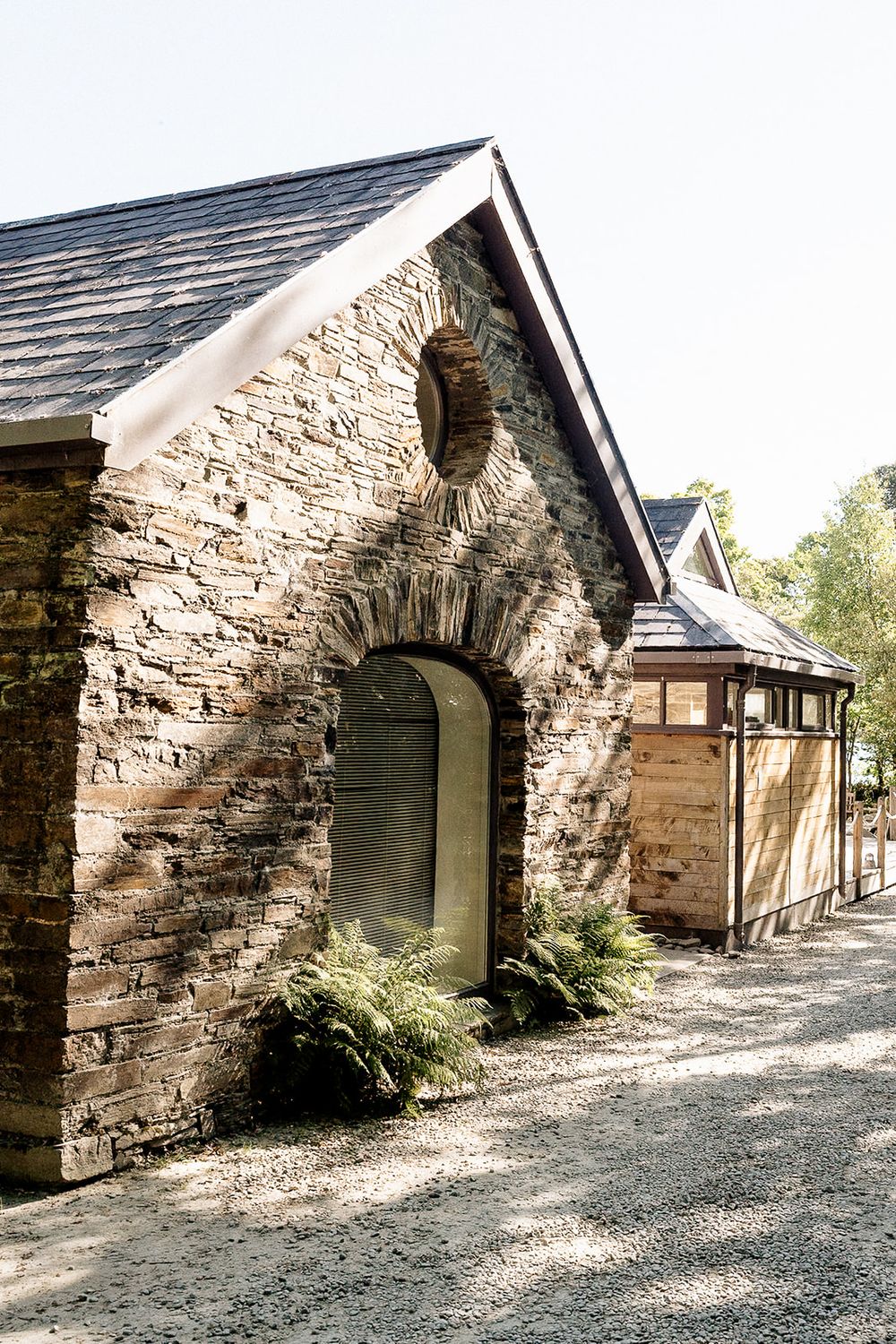A rustic stone building entrance with an arched doorway and ferns growing alongside the weathered facade.