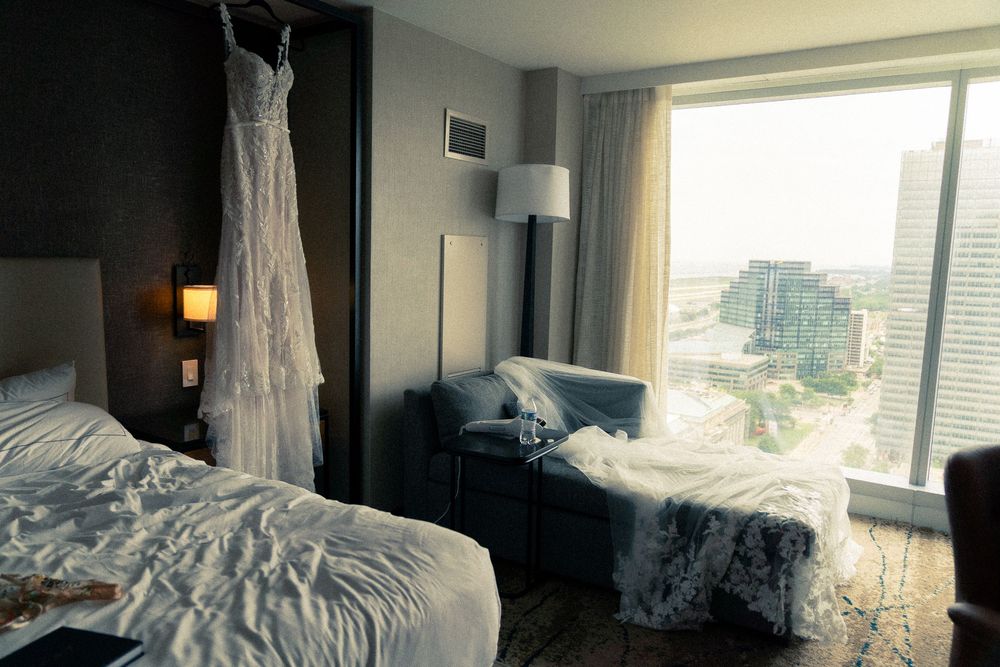 A sunlit bedroom interior with rumpled white bedding and city views through large windows during different times of day.