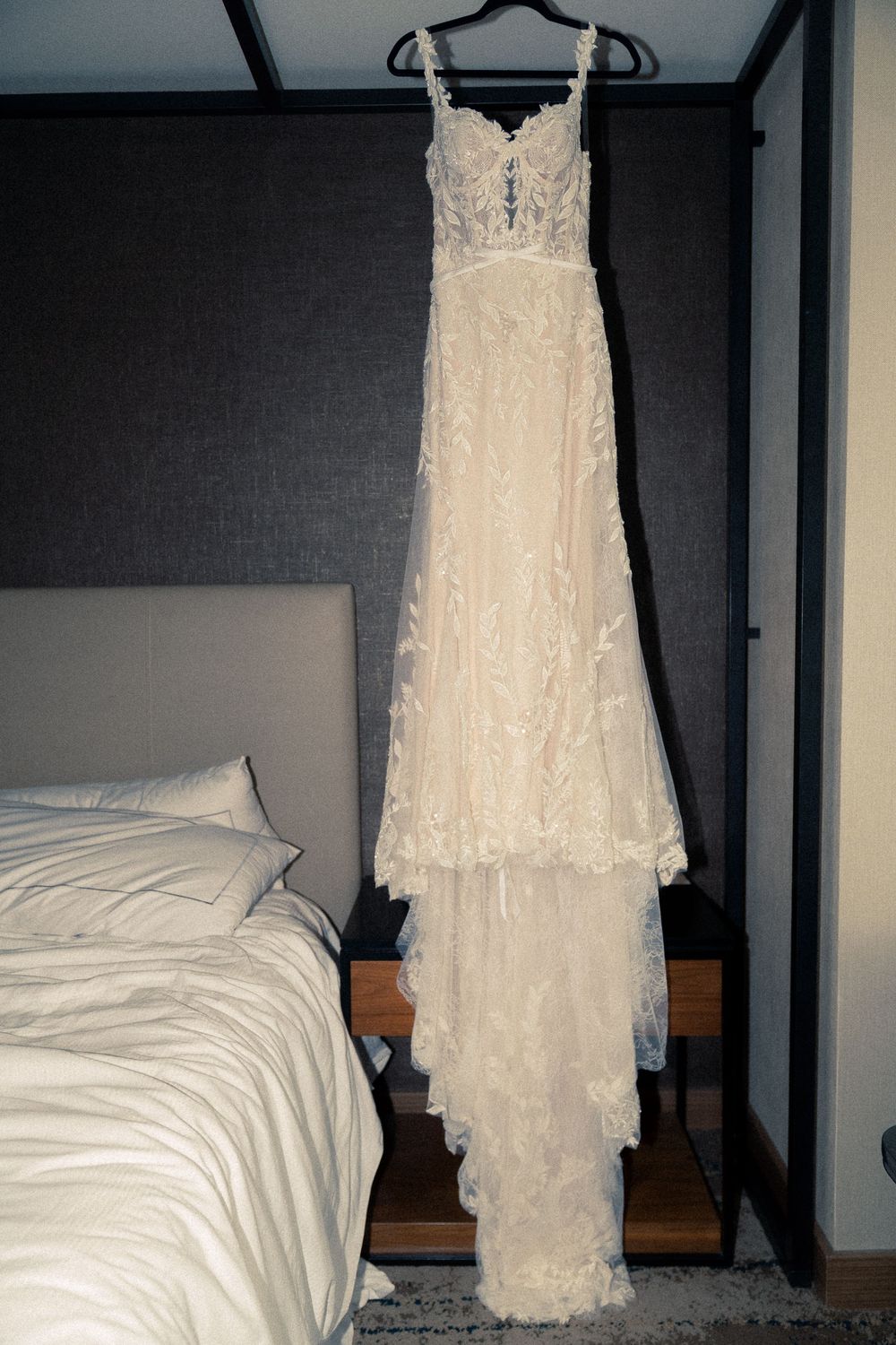 A white lace wedding dress hangs on a black hanger against a white wall with a bed visible in the background.