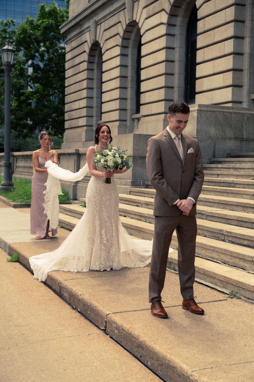 Bride in lace gown approaches groom for first look outside historic stone building, bridesmaid holding train.