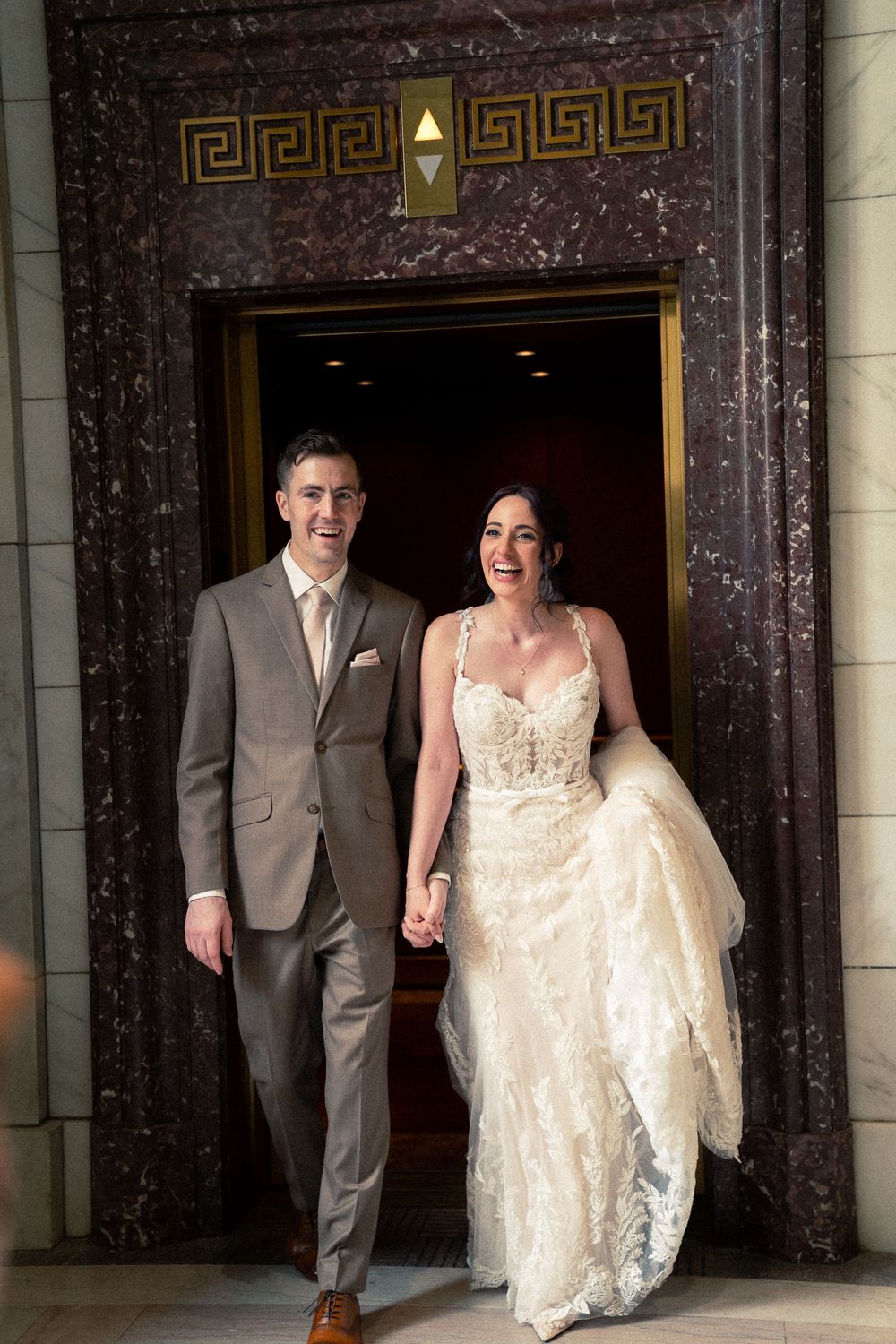 A couple in wedding attire walks through an elegant marble doorway with Greek key pattern detailing.