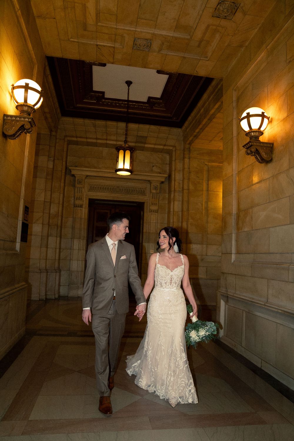 A couple walks down an elegant hallway with stone walls and vintage lighting fixtures on their wedding day.