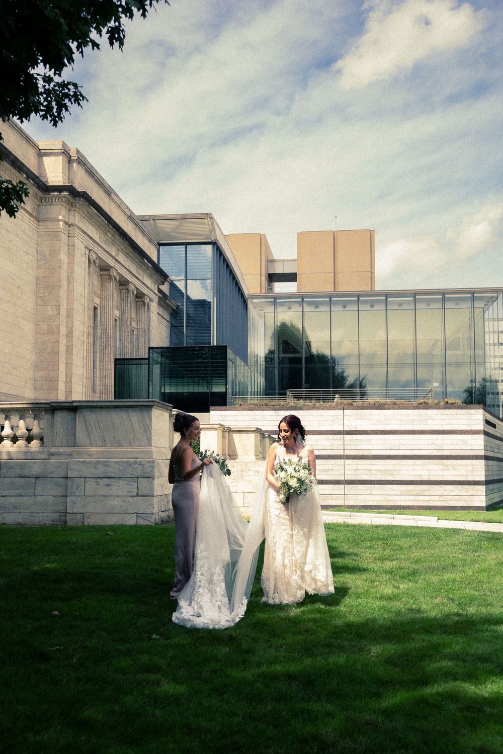 A couple in wedding attire sits on a grassy lawn outside a modern building with glass windows and concrete walls.