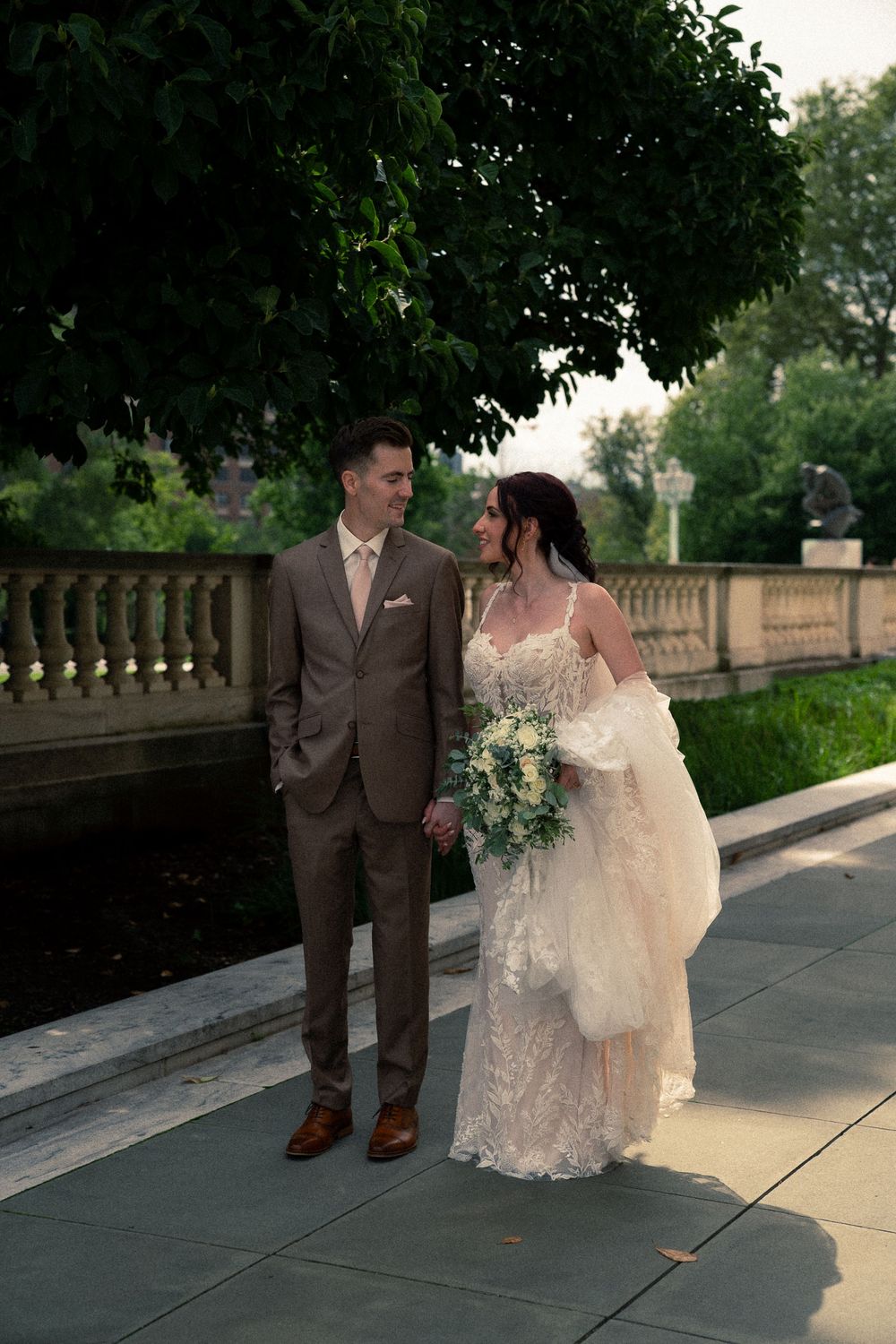 A couple in wedding attire walks along a stone path during an outdoor photo session at dusk.