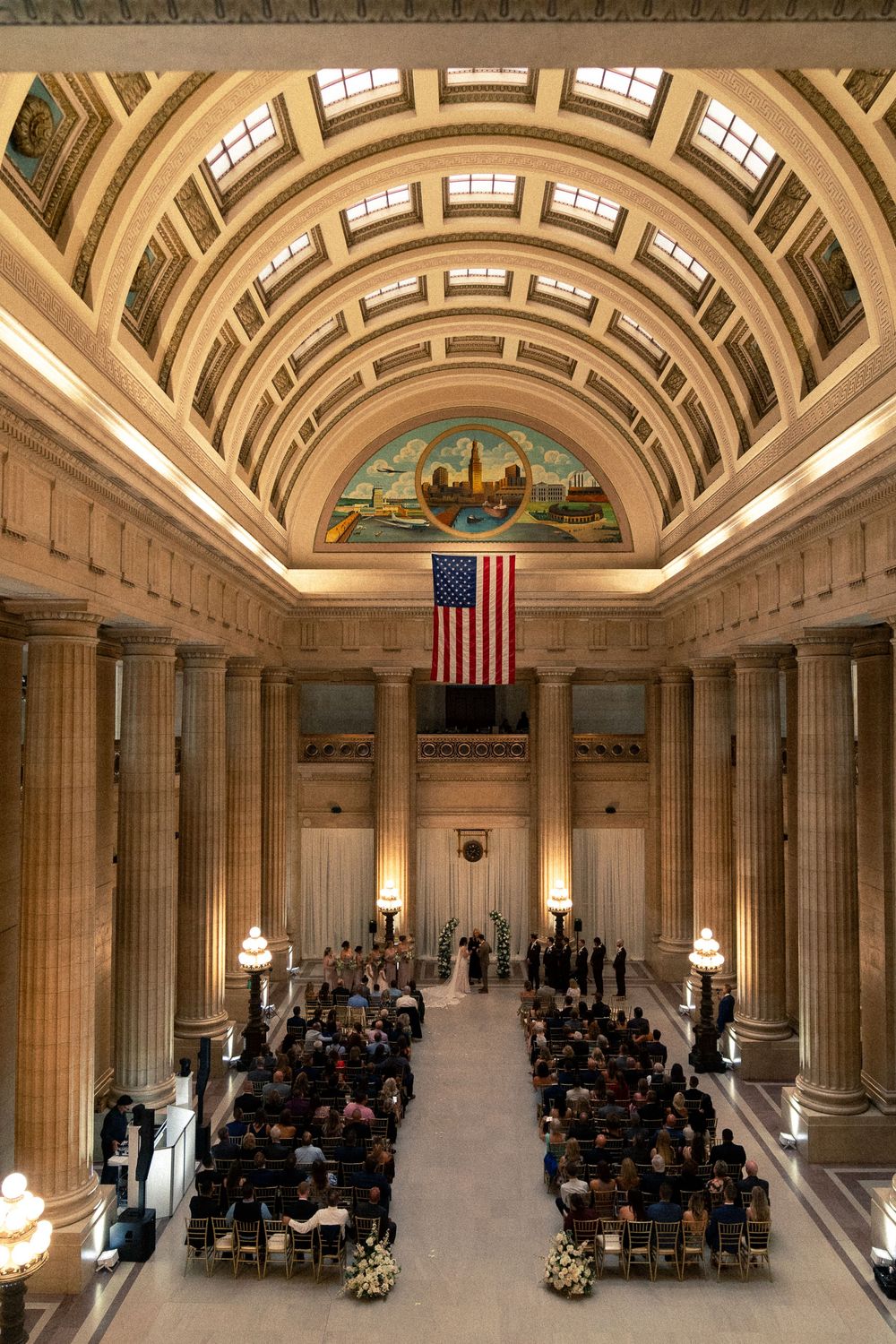Grand ceremony taking place in historic courthouse hall with arched ceiling, columns and American flag hanging prominently.