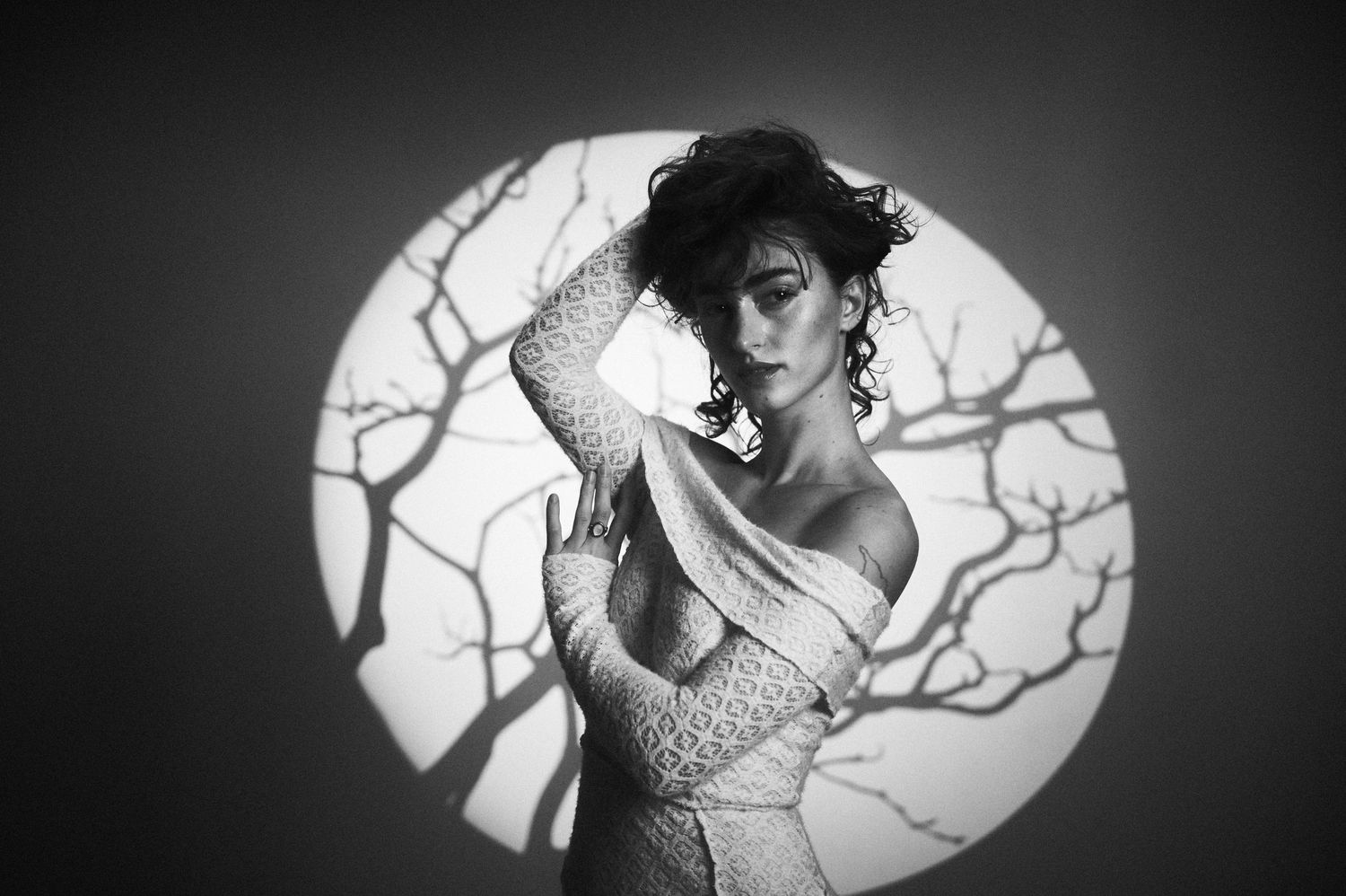 Dramatic black and white portrait with a circular vignette showing figure in lace against silhouetted tree branches.