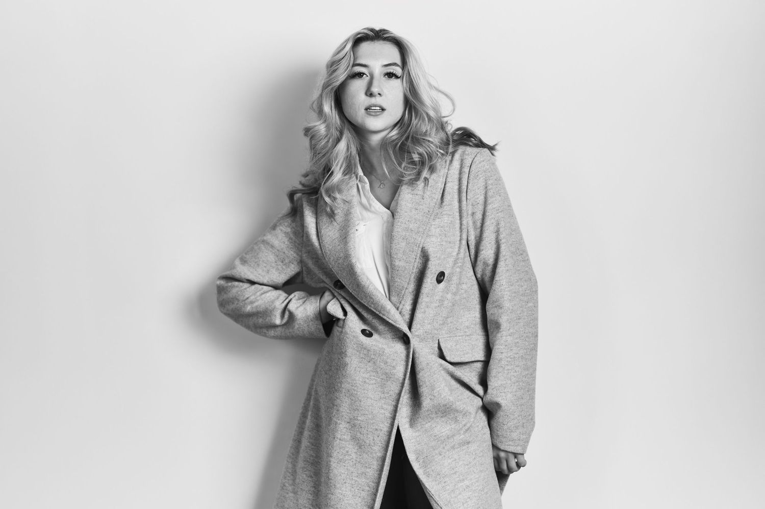 Fashion model wearing a long gray wool coat poses against white background in black and white photograph.