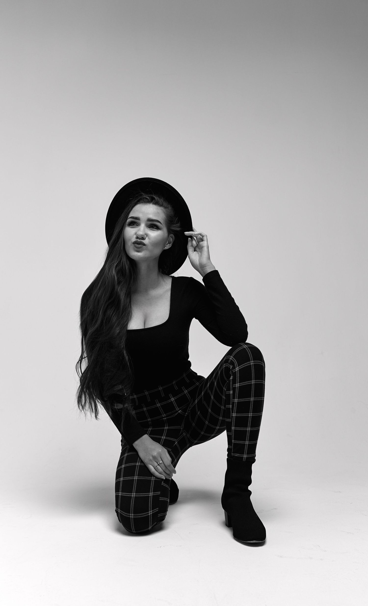Artistic black and white portrait of someone in a hat, black top, plaid pants and boots crouching against white backdrop.
