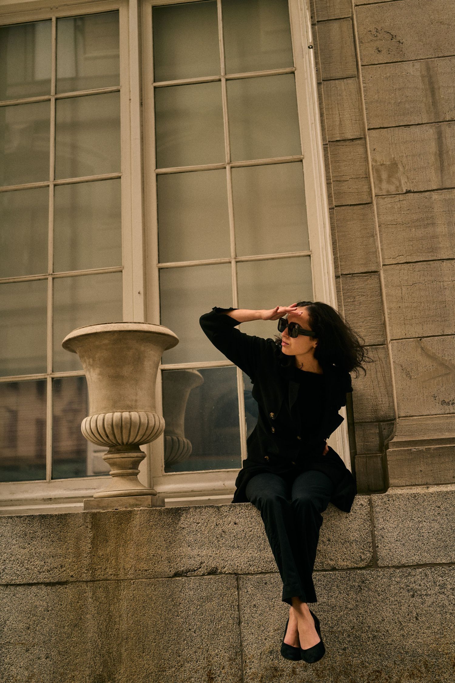 A person in black attire and sunglasses sits on a stone windowsill next to a classical urn in moody lighting.
