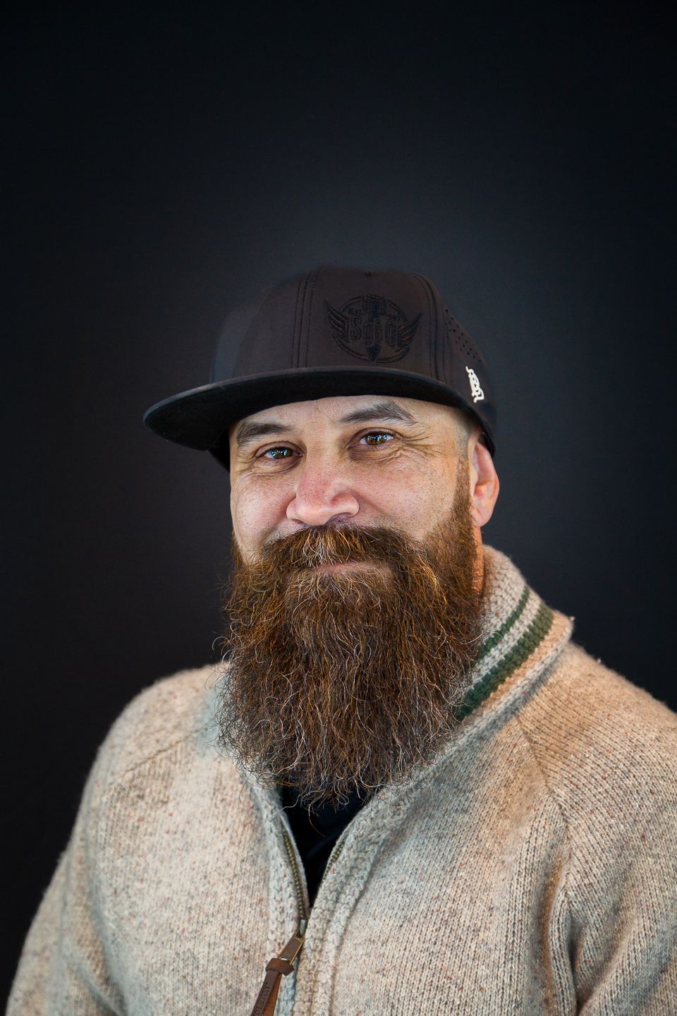 Portrait of someone wearing a baseball cap and cable knit sweater with a long reddish beard against dark background.
