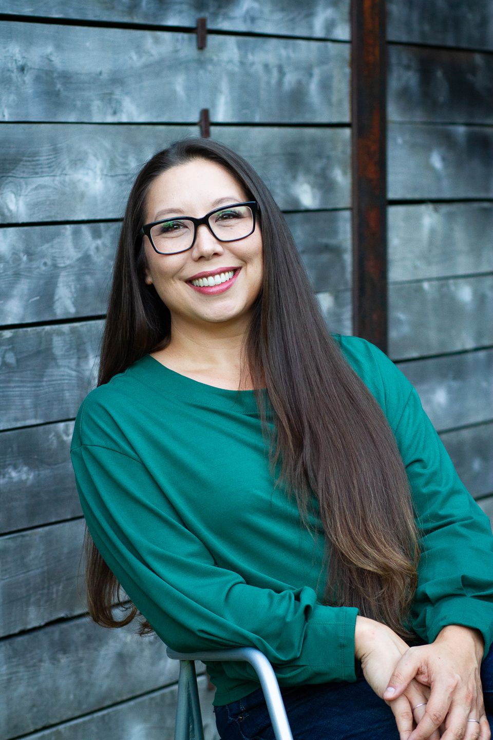 A warm smile and dark flowing hair complement stylish glasses and an emerald green sweater against a wood panel wall.